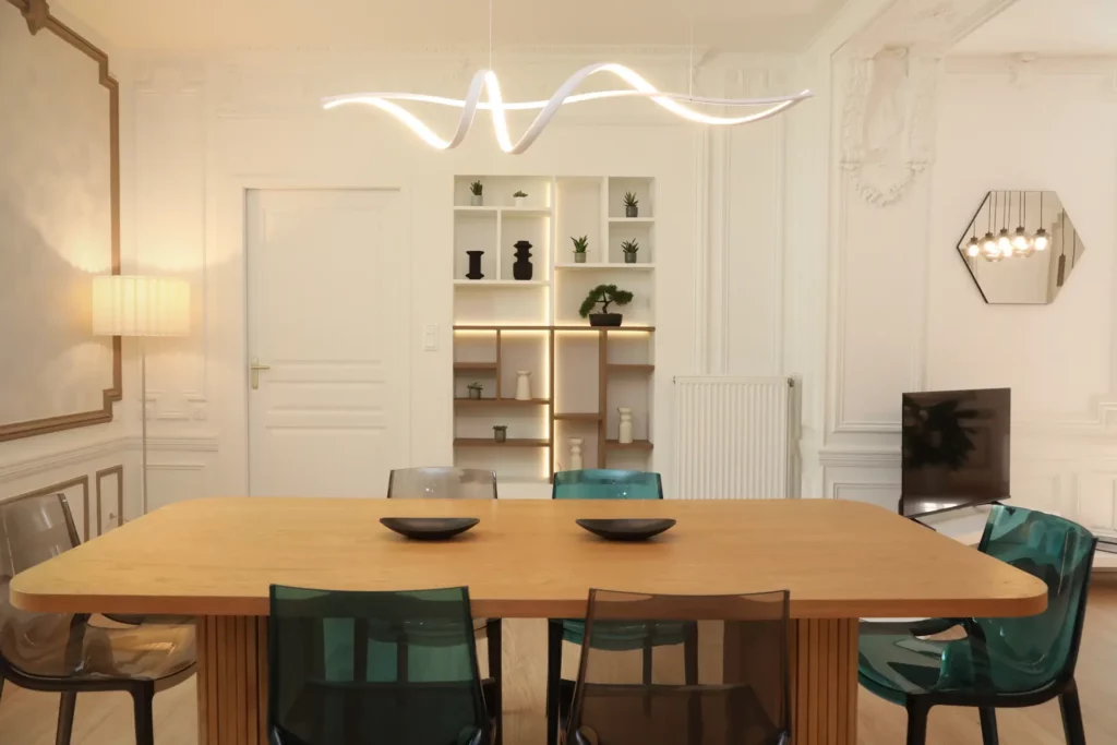 Salle à manger design dans un grand T5 à Chantilly par Ilot Archi avec grande table en bois, chaises colorées et niche décorative éclairée.