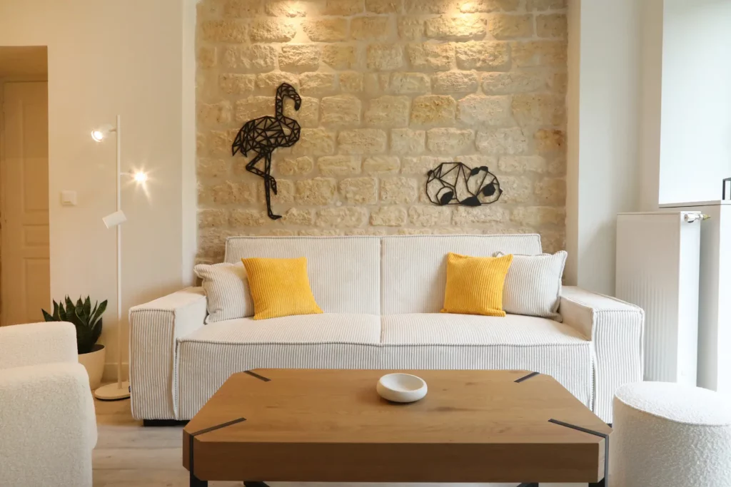 Salon chaleureux aménagé par Ilot Archi à Chantilly avec canapé blanc, coussins jaunes et décoration murale en métal sur mur en pierres.