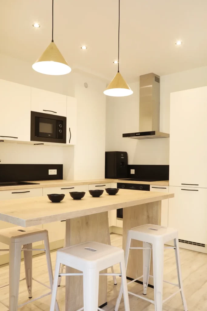 Agencement d'une cuisine ouverte blanche et bois avec mange-debout et suspensions design dans un T2 par Ilot Archi.