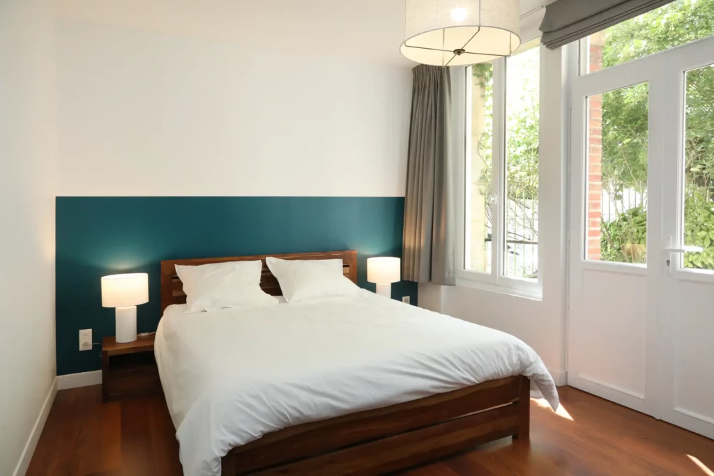 Agencement d'une chambre lumineuse à Chantilly par Ilot Archi avec soubassement bleu pétrole, lit en bois massif et baies vitrées.