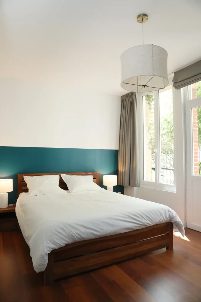 Décoration d'une chambre moderne rénovée par Ilot Archi avec mur peint bleu canard, parquet chaleureux et grand luminaire suspension.