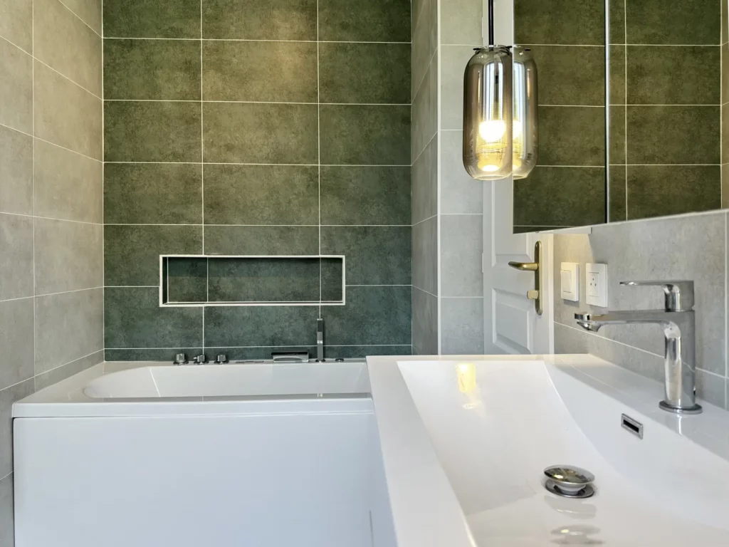Salle de bains rénovée par Ilot Archi avec baignoire, carrelage vert sauge, niche encastrée et meuble double vasque.